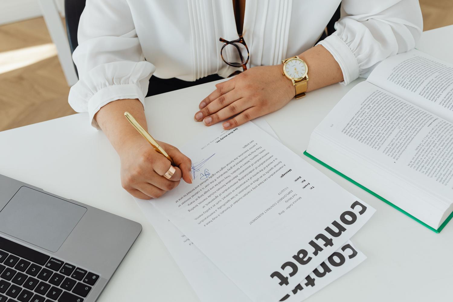 Mujer profesional firmando un contrato mercantil en su escritorio con un portátil y libros legales Mujer profesional firmando un contrato mercantil en su escritorio con un portátil y libros legales
