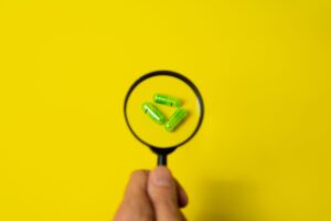 Persona sosteniendo una lupa sobre cápsulas verdes sobre un fondo amarillo brillante, representando el análisis detallado de productos farmacéuticos en el contexto del marketing farmacéutico