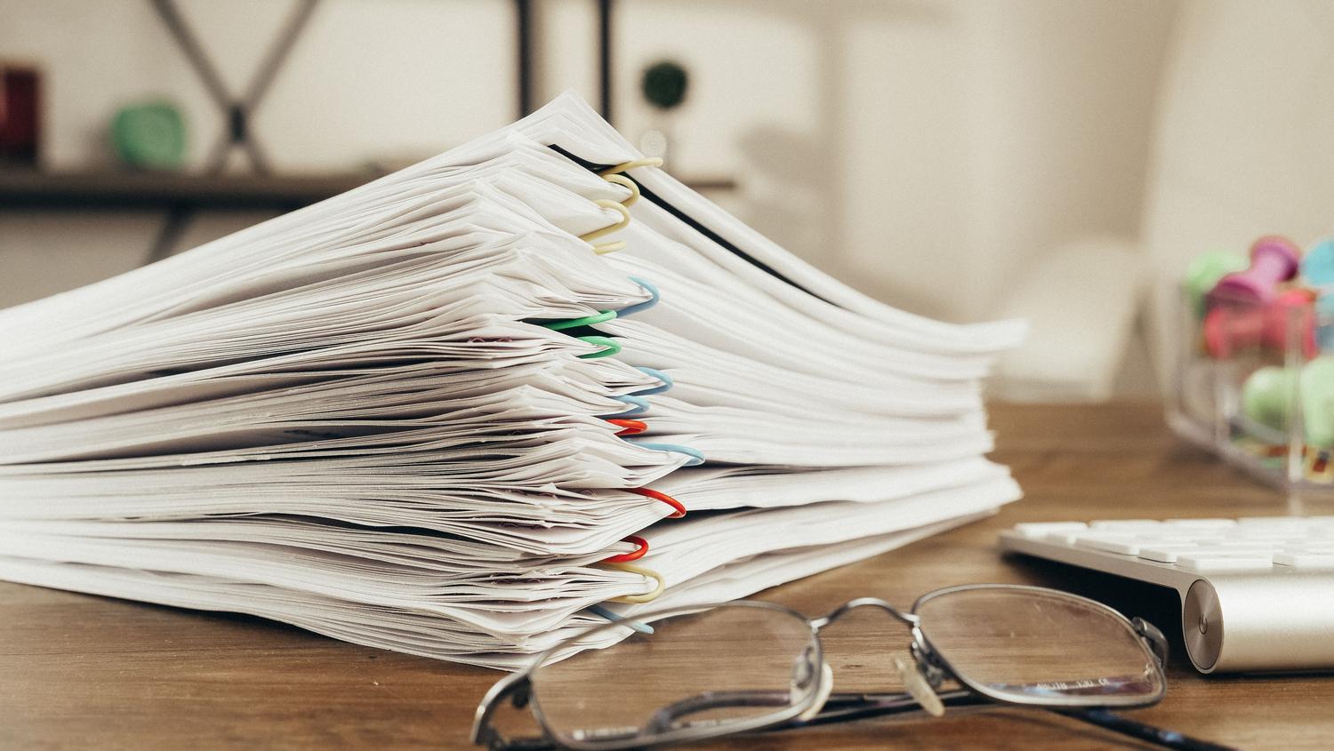 Primer plano de una pila de documentos administrativos con gafas sobre la mesa, ilustrando la burocracia y el volumen de papelería requerida. Primer plano de una pila de documentos administrativos con gafas sobre la mesa, ilustrando la burocracia y el volumen de papelería requerida.