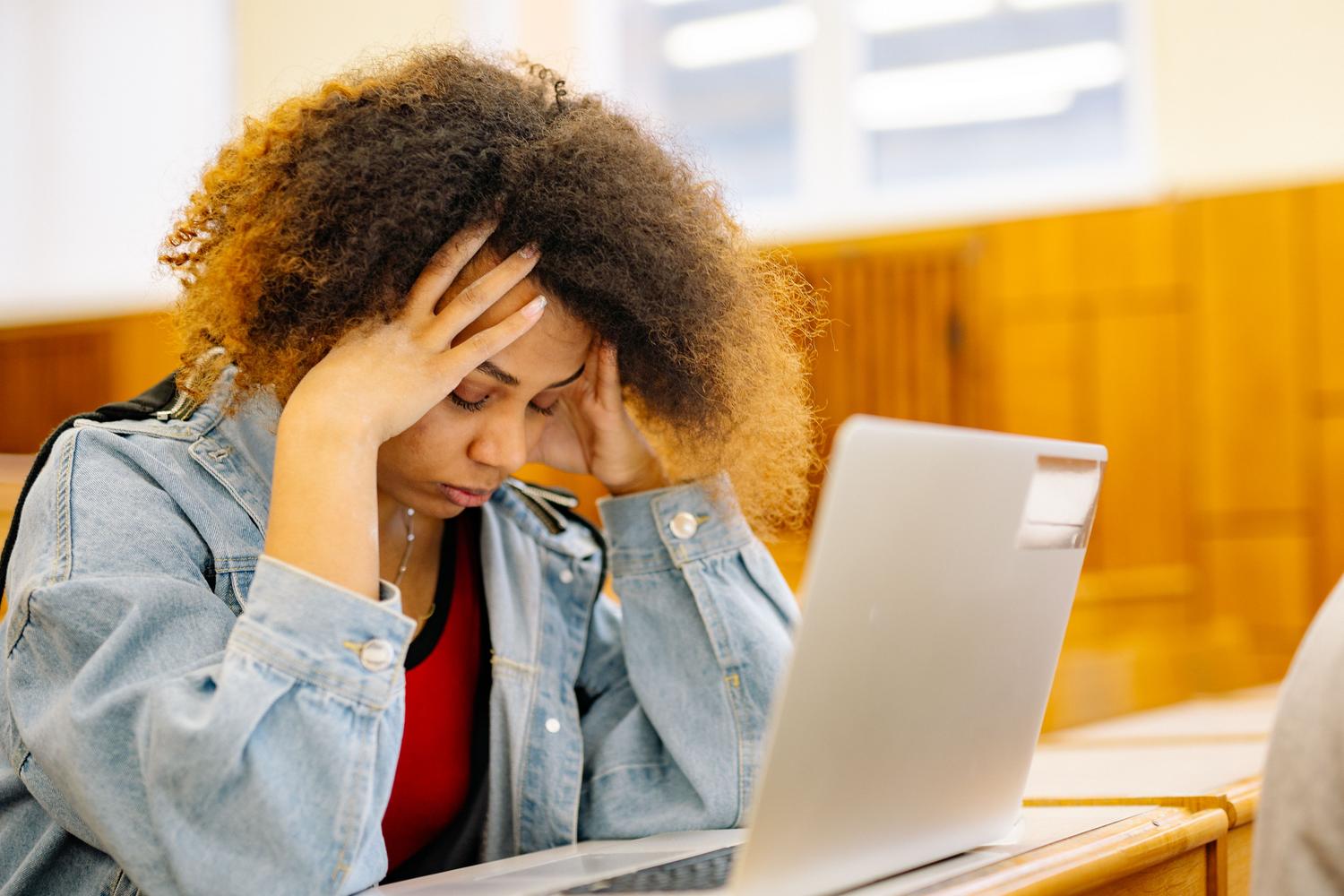 Joven estudiante con dolor de cabeza y expresión de agotamiento mientras utiliza un ordenador portátil para estudiar. Joven estudiante con dolor de cabeza y expresión de agotamiento mientras utiliza un ordenador portátil para estudiar.