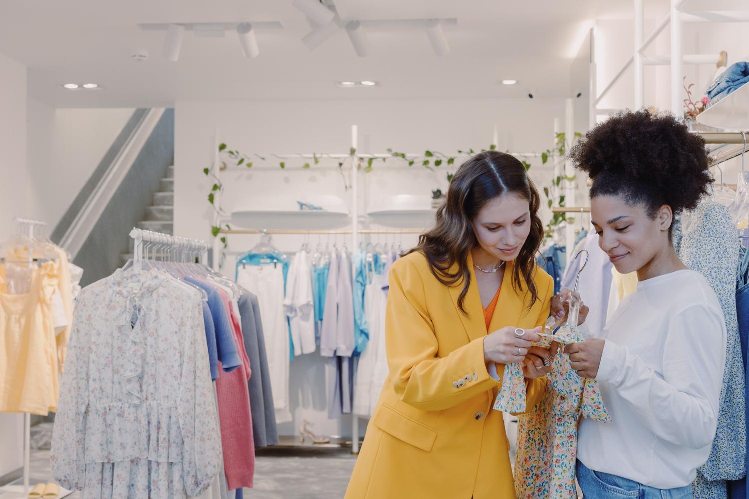 Experiencia de venta en tiendas físicas modernas Dos mujeres explorando ropa en una boutique moderna y elegante, ilustrando la experiencia de venta en tiendas físicas.