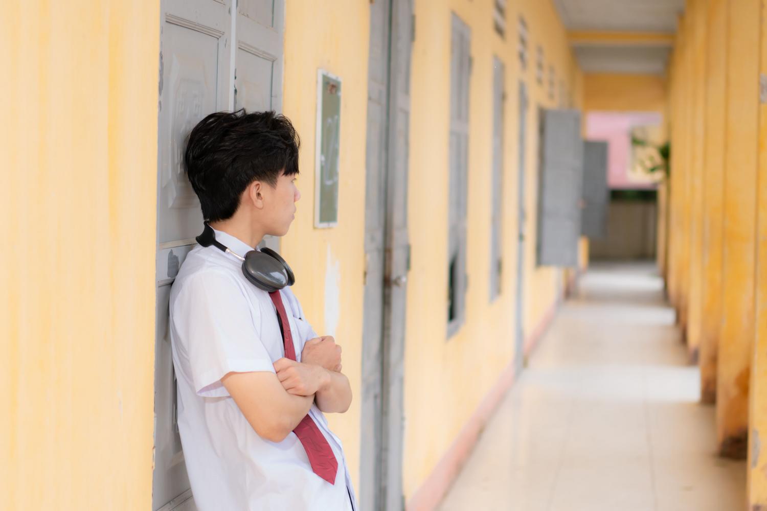 Un estudiante adolescente apoyado en la pared de un pasillo escolar, pensativo y distante, ilustrando la desconexión emocional con el centro educativo. Un estudiante adolescente apoyado en la pared de un pasillo escolar, pensativo y distante, ilustrando la desconexión emocional con el centro educativo.
