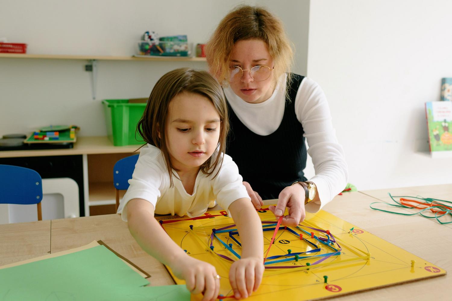 Terapeuta ocupacional guiando a una niña en una actividad de motricidad fina con un tablero geométrico en un aula. Terapeuta ocupacional guiando a una niña en una actividad de motricidad fina con un tablero geométrico en un aula.