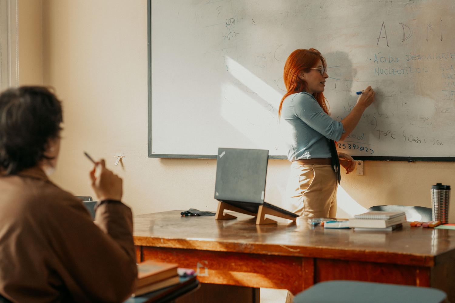 Profesora universitaria explicando conceptos en la pizarra apoyada por un ordenador portátil, destacando el uso didáctico de la tecnología en el aula. Profesora universitaria explicando conceptos en la pizarra apoyada por un ordenador portátil, destacando el uso didáctico de la tecnología en el aula.