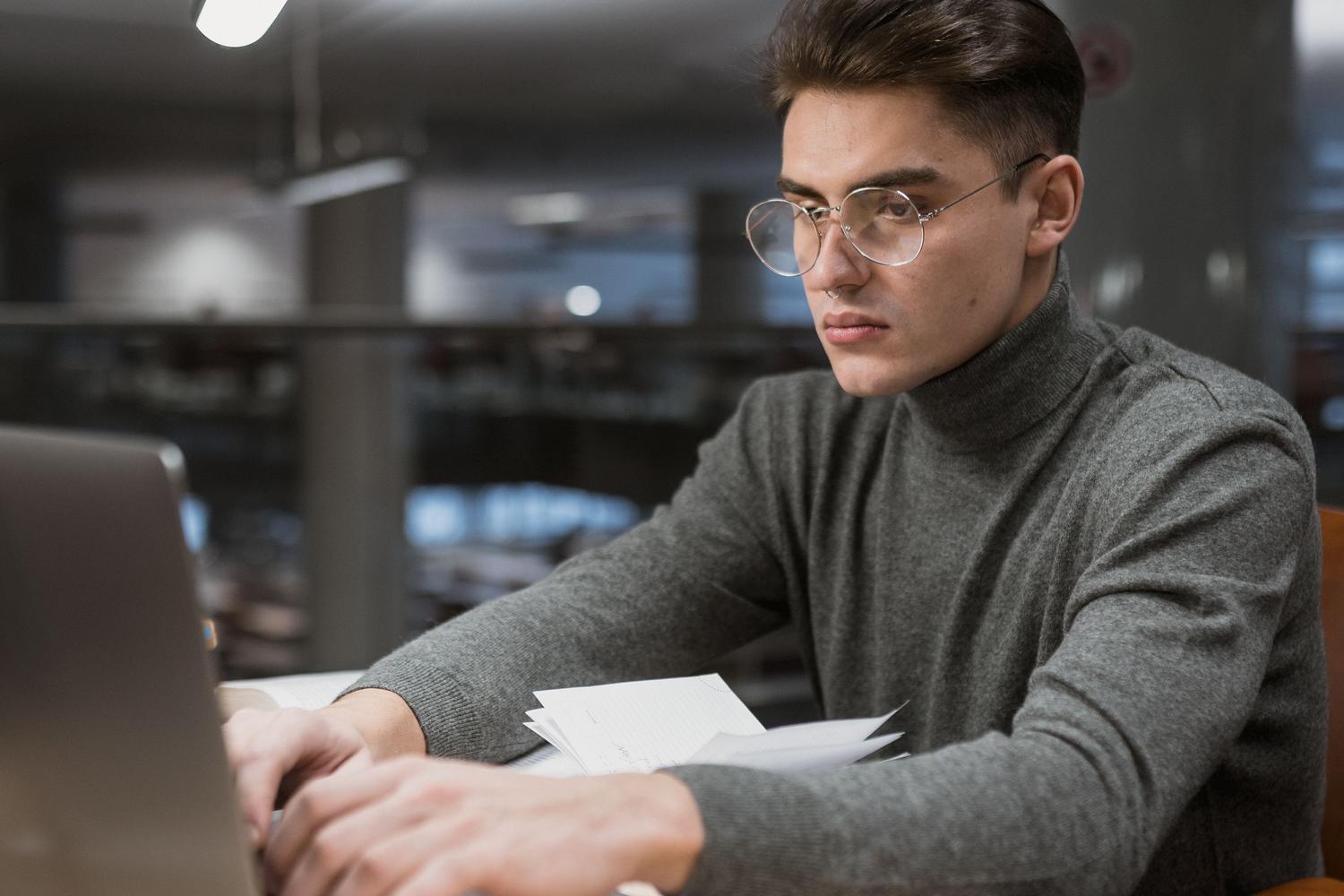 Estudiante universitario concentrado estudiando con un ordenador portátil en una biblioteca moderna. Estudiante universitario concentrado estudiando con un ordenador portátil en una biblioteca moderna.