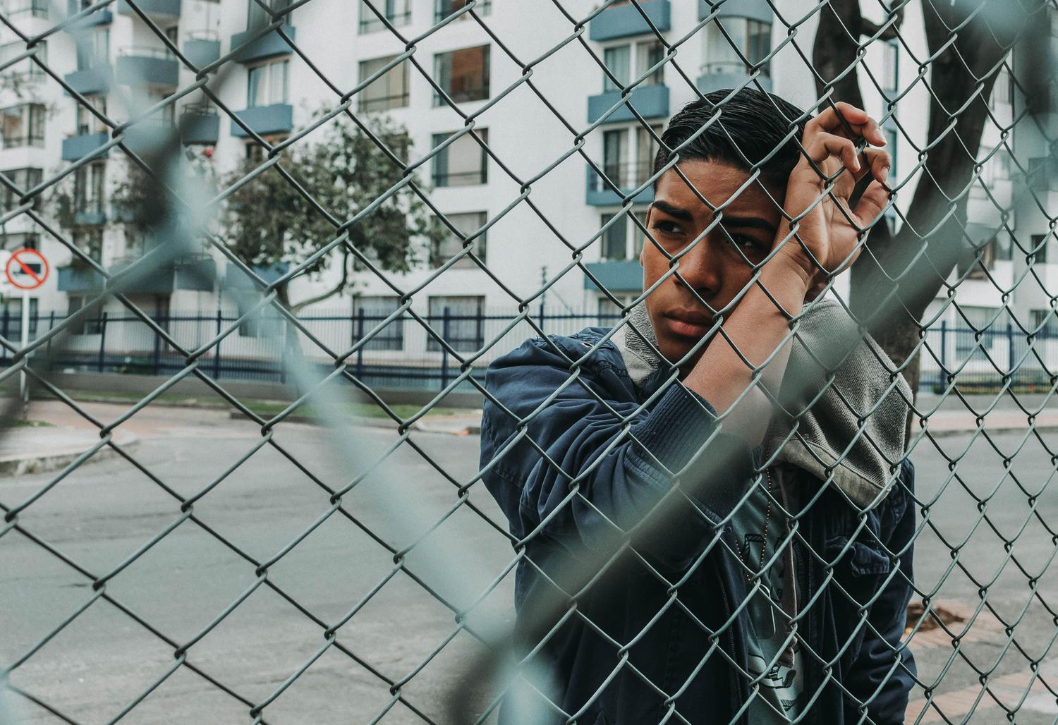Joven pensativo apoyado en una valla metálica en un entorno urbano, representando los límites del internamiento y la reflexión personal. Joven pensativo apoyado en una valla metálica en un entorno urbano, representando los límites del internamiento y la reflexión personal.