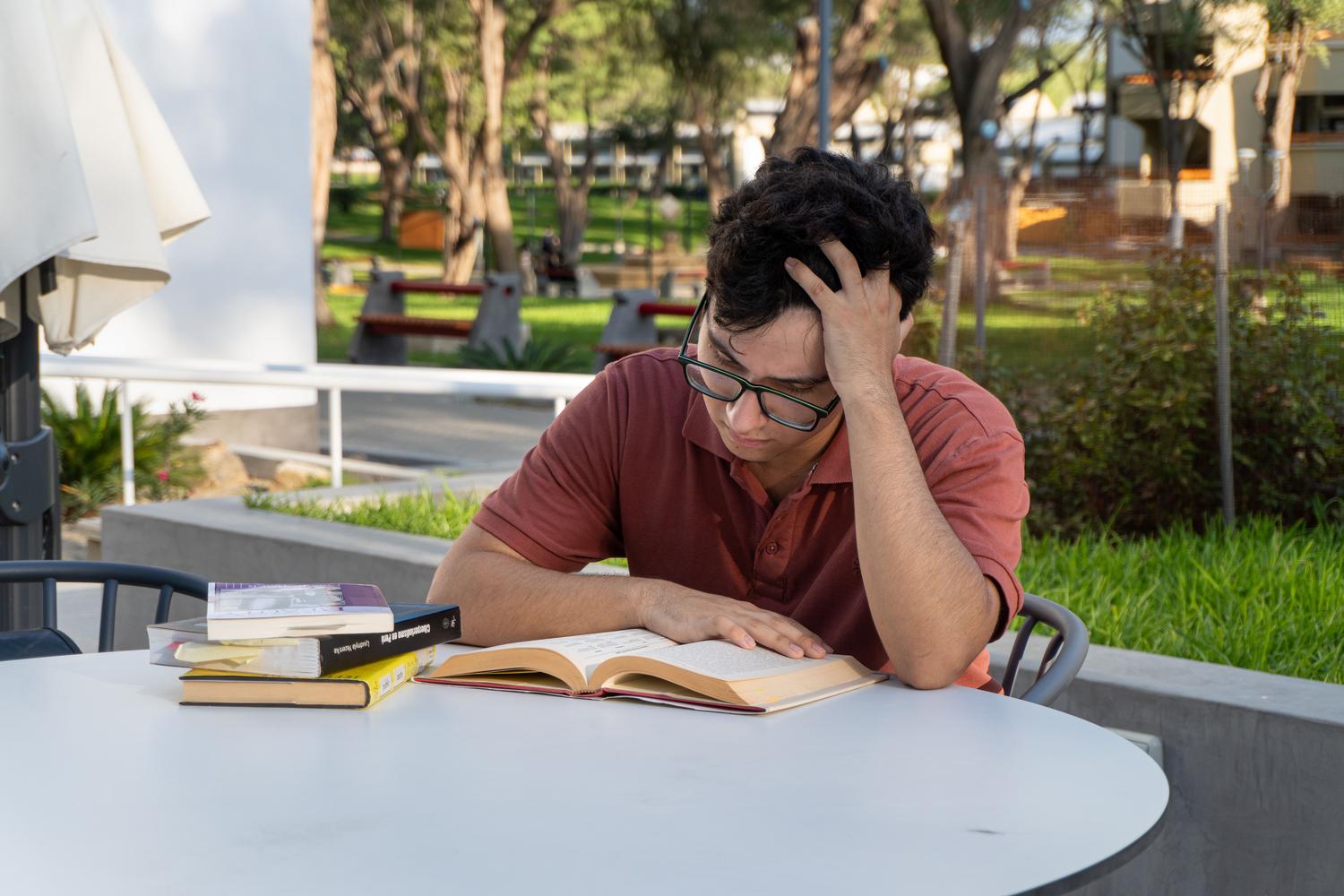 Joven estudiante concentrado estudiando libros al aire libre en el campus, representando el esfuerzo académico necesario para alcanzar las notas de corte. Joven estudiante concentrado estudiando libros al aire libre en el campus, representando el esfuerzo académico necesario para alcanzar las notas de corte.