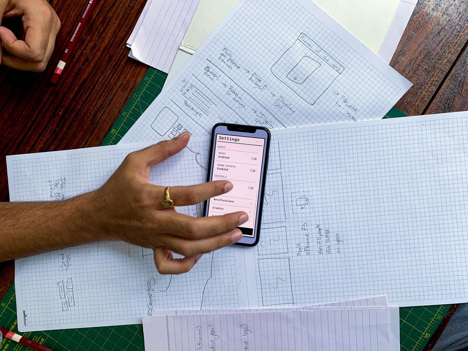 Vista cenital de un espacio de trabajo con bocetos de wireframes hechos a mano y un smartphone mostrando la interfaz de una aplicación, representando el paso de la idea al prototipo. Vista cenital de un espacio de trabajo con bocetos de wireframes hechos a mano y un smartphone mostrando la interfaz de una aplicación, representando el paso de la idea al prototipo.