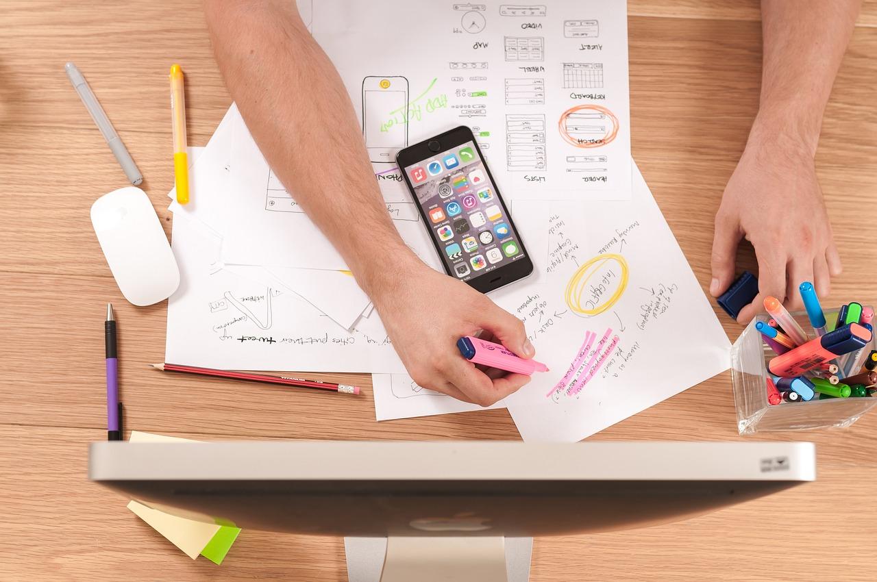 Mesa de diseño con bocetos de aplicaciones móviles y un teléfono inteligente con una interfaz navegable, ejemplificando la creación de productos mediante plataformas no-code. Mesa de diseño con bocetos de aplicaciones móviles y un teléfono inteligente con una interfaz navegable, ejemplificando la creación de productos mediante plataformas no-code.
