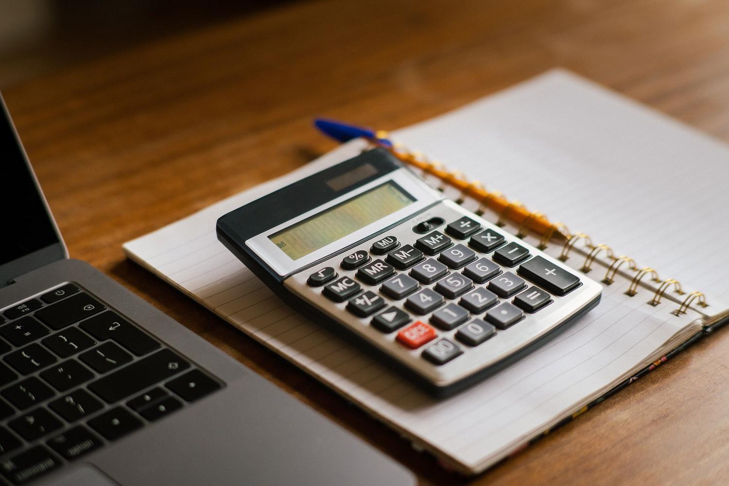Calculadora y ordenador portátil sobre una mesa de madera, representando el cálculo de cuotas e impuestos. Calculadora y ordenador portátil sobre una mesa de madera, representando el cálculo de cuotas e impuestos.