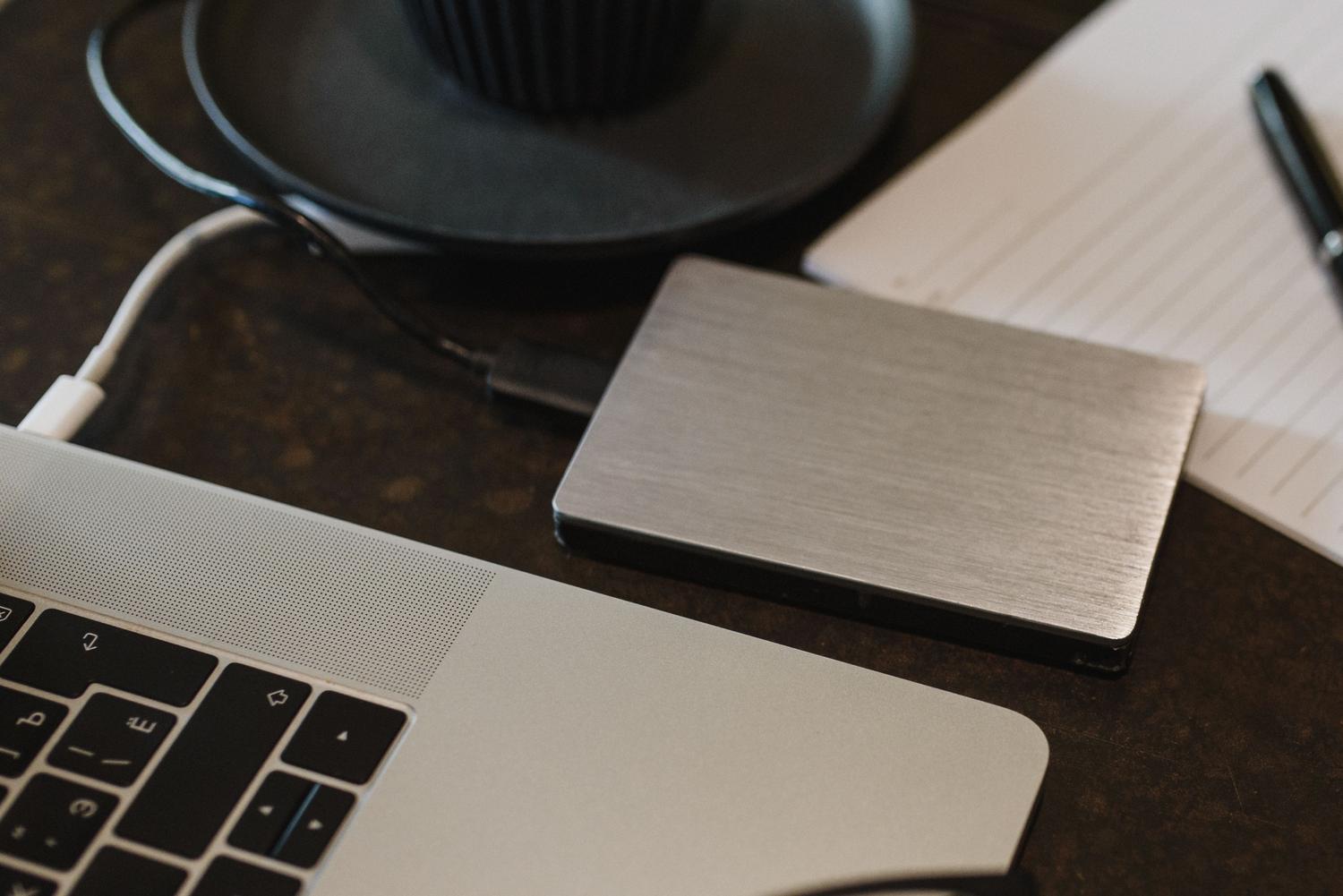 Primer plano de un disco duro externo conectado a un ordenador portátil en un escritorio, representando la gestión de copias de seguridad de datos. Primer plano de un disco duro externo conectado a un ordenador portátil en un escritorio, representando la gestión de copias de seguridad de datos.