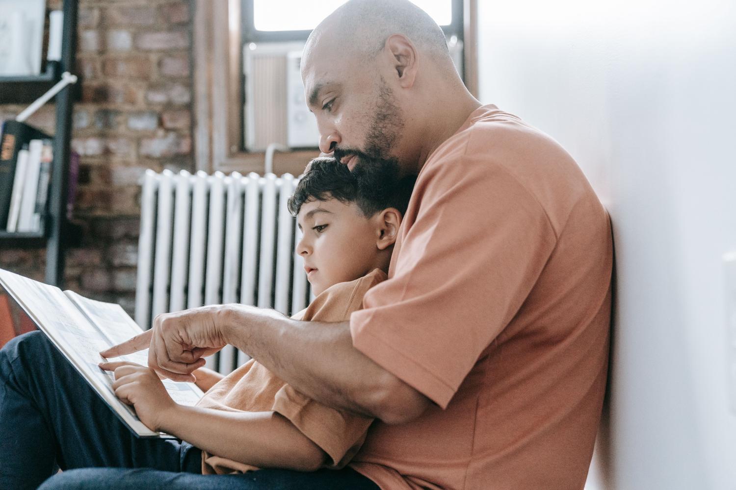 Padre enseñando a su hijo a leer en un ambiente hogareño, representando la alianza vital entre la familia y la escuela para fomentar el pensamiento. Padre enseñando a su hijo a leer en un ambiente hogareño, representando la alianza vital entre la familia y la escuela para fomentar el pensamiento.