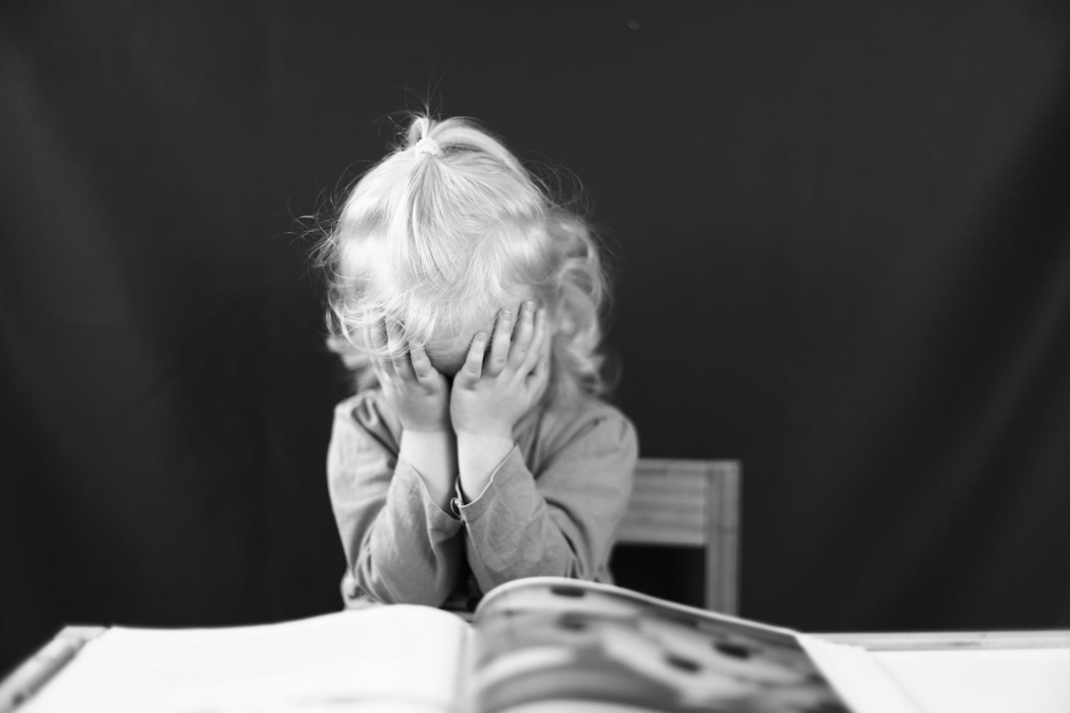 Niña pequeña en blanco y negro cubriendo su rostro con las manos frente a un libro, transmitiendo desesperación y presión. Niña pequeña en blanco y negro cubriendo su rostro con las manos frente a un libro, transmitiendo desesperación y presión.