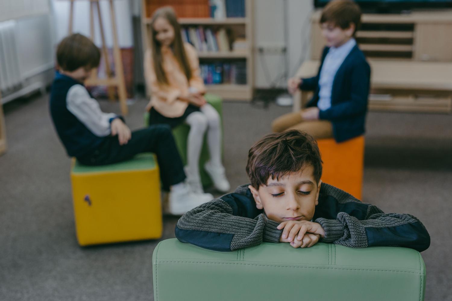 Niño pequeño con mirada triste y retraída en un aula, contrastando con otros niños que juegan al fondo, ilustrando el aislamiento. Niño pequeño con mirada triste y retraída en un aula, contrastando con otros niños que juegan al fondo, ilustrando el aislamiento.