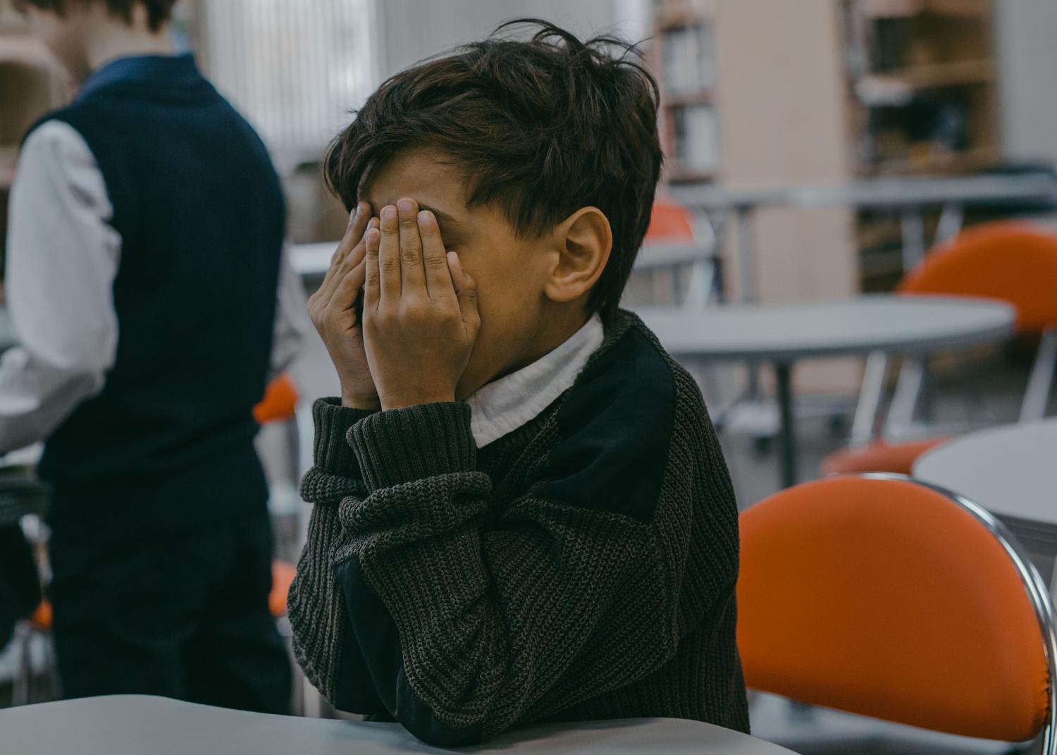 Niño con uniforme escolar cubriendo su cara con las manos sobre una mesa blanca, reflejando agotamiento emocional y retraimiento. Niño con uniforme escolar cubriendo su cara con las manos sobre una mesa blanca, reflejando agotamiento emocional y retraimiento.