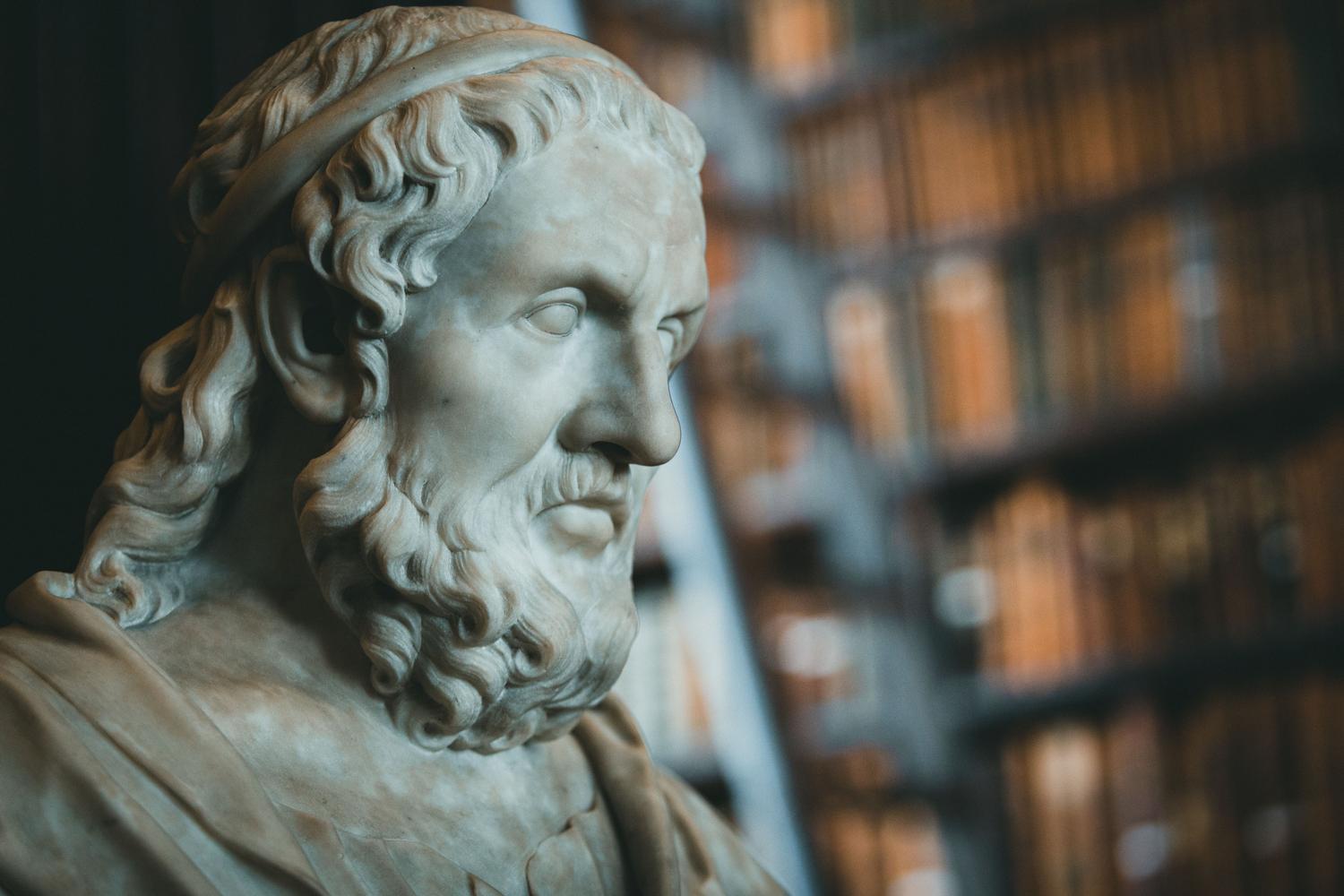 Estatua de mármol clásica frente a una estantería de libros, representando la unión entre la filosofía antigua y el estudio académico. Estatua de mármol clásica frente a una estantería de libros, representando la unión entre la filosofía antigua y el estudio académico.