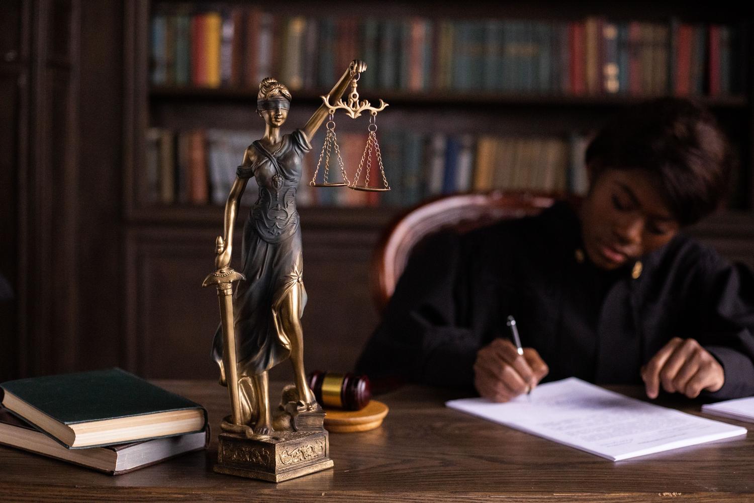 Profesional del derecho redactando documentos junto a la estatua de la Dama de la Justicia y libros jurídicos. Profesional del derecho redactando documentos junto a la estatua de la Dama de la Justicia y libros jurídicos.