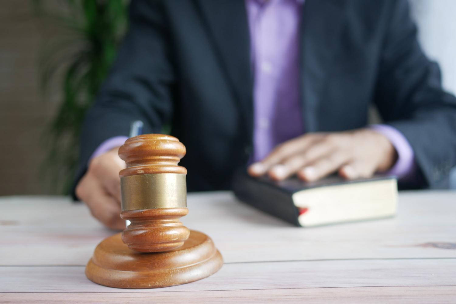 Mazo de juez y libro legal en primer plano con un profesional escribiendo al fondo, representando la redacción de fundamentos jurídicos. Mazo de juez y libro legal en primer plano con un profesional escribiendo al fondo, representando la redacción de fundamentos jurídicos.
