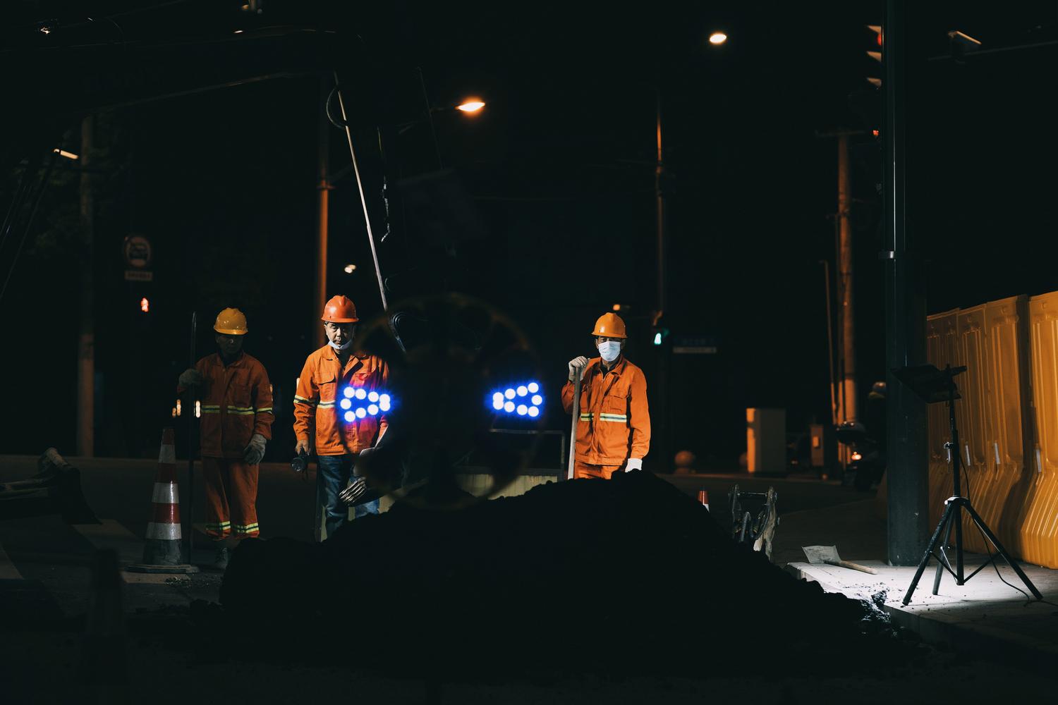 Trabajadores viales con ropa de alta visibilidad y cascos realizando labores de mantenimiento nocturno bajo luces artificiales. Trabajadores viales con ropa de alta visibilidad y cascos realizando labores de mantenimiento nocturno bajo luces artificiales.