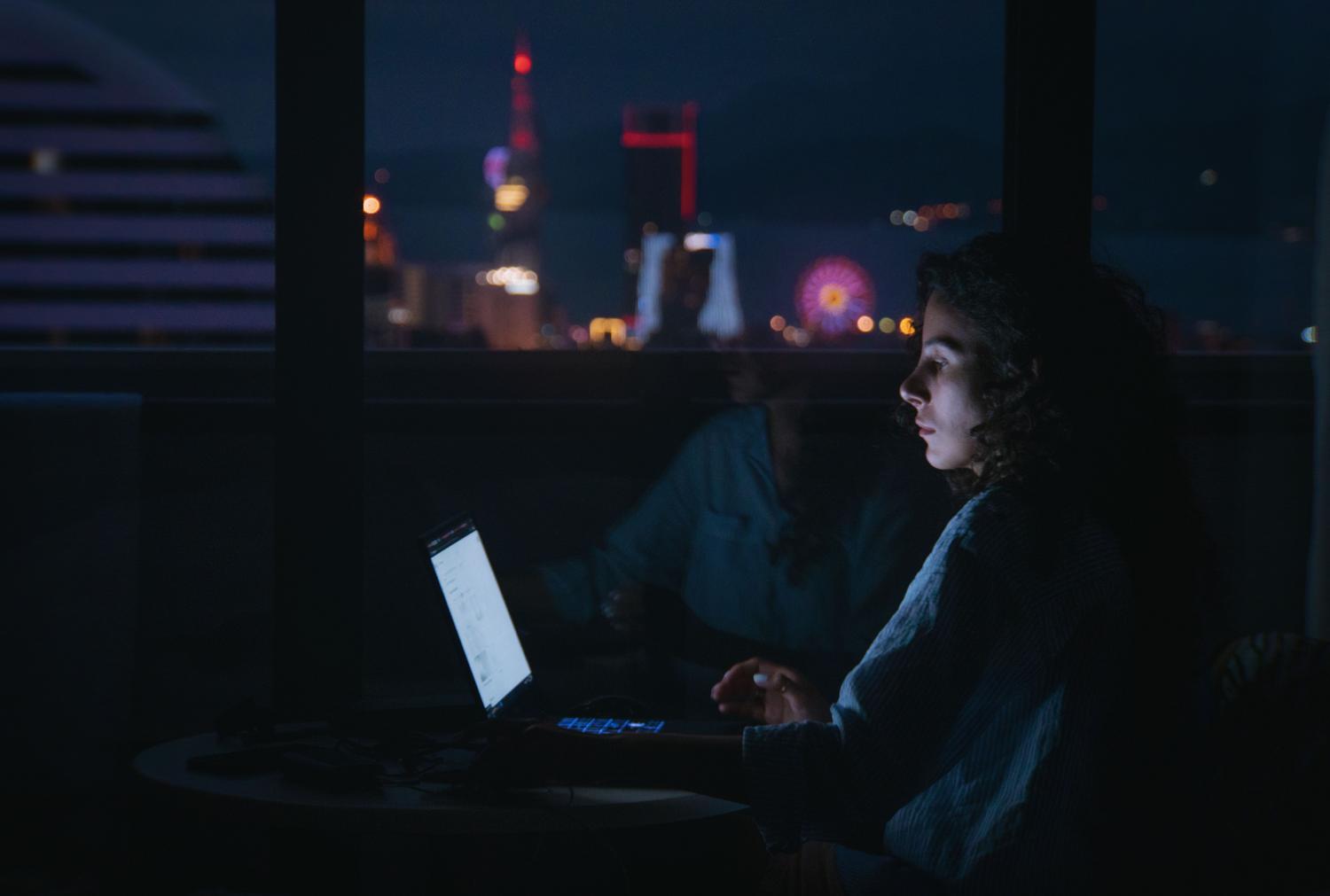 Mujer concentrada trabajando con su portátil en una habitación oscura con las luces de la ciudad visibles al fondo. Mujer concentrada trabajando con su portátil en una habitación oscura con las luces de la ciudad visibles al fondo.