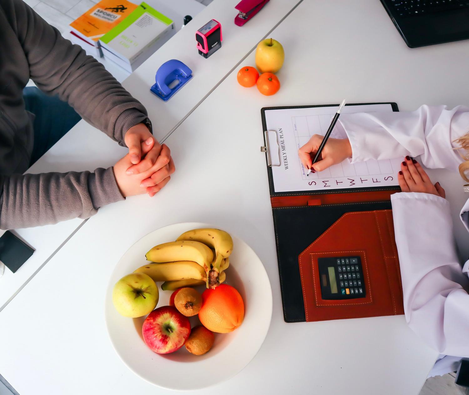 Vista superior de una dietista diseñando un plan nutricional personalizado con frutas frescas sobre la mesa. Vista superior de una dietista diseñando un plan nutricional personalizado con frutas frescas sobre la mesa.
