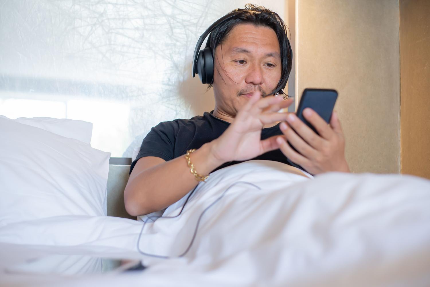 Hombre joven relajado en su cama utilizando auriculares y un smartphone para escuchar un podcast de inmersión lingüística. Hombre joven relajado en su cama utilizando auriculares y un smartphone para escuchar un podcast de inmersión lingüística.