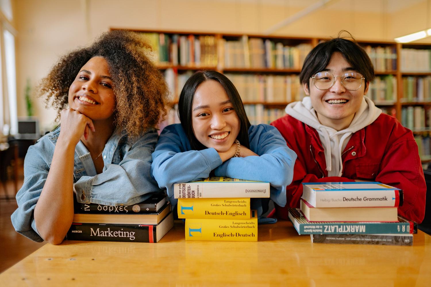 Tres estudiantes universitarios sonrientes en una biblioteca estudiando con diccionarios y libros de gramática alemana. Tres estudiantes universitarios sonrientes en una biblioteca estudiando con diccionarios y libros de gramática alemana.