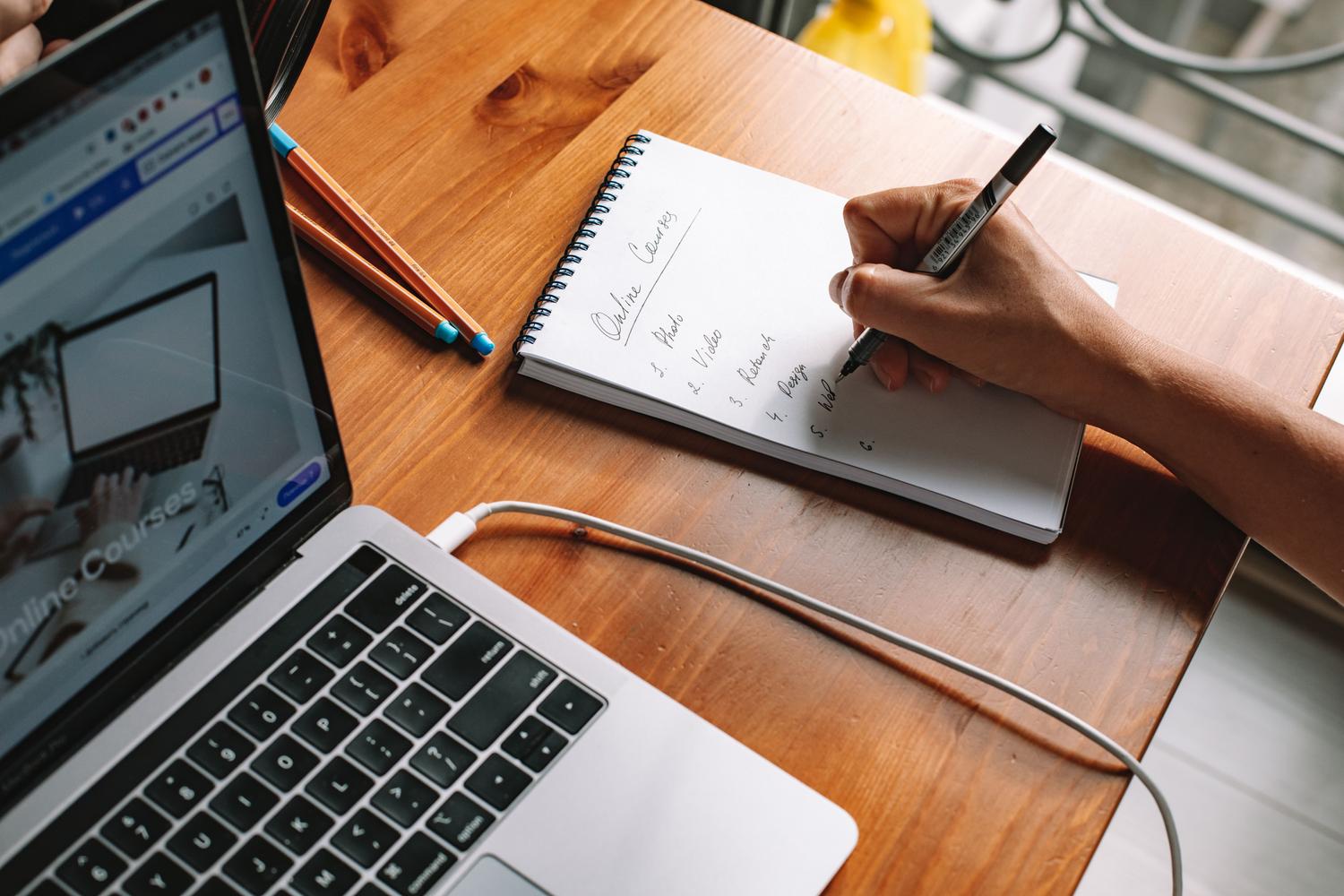 Persona tomando notas en un cuaderno mientras investiga cursos en línea en su computadora portátil. Persona tomando notas en un cuaderno mientras investiga cursos en línea en su computadora portátil.