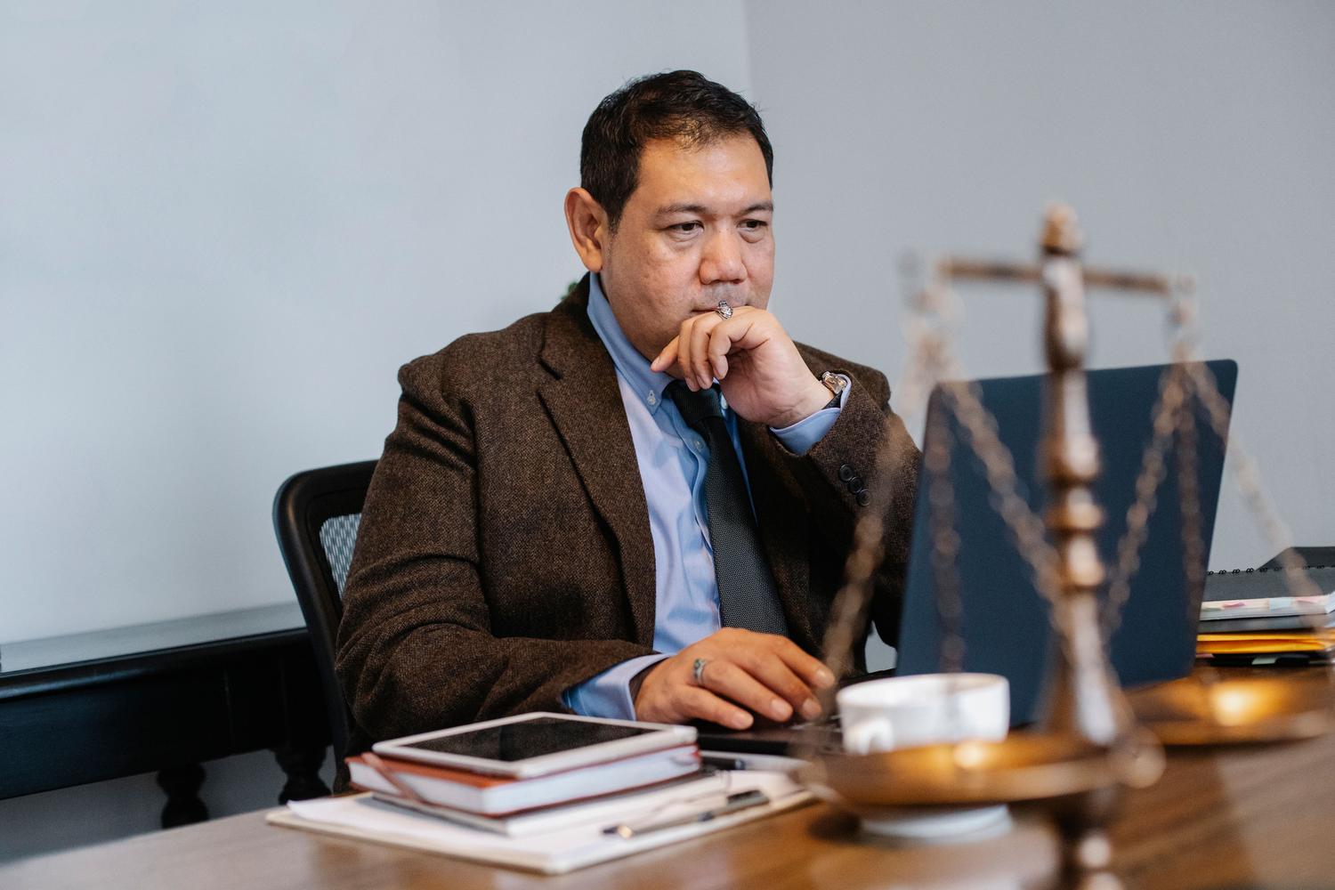 Abogado analizando normativas tecnológicas en su portátil, rodeado de libros de derecho y la balanza de la justicia. Abogado analizando normativas tecnológicas en su portátil, rodeado de libros de derecho y la balanza de la justicia.