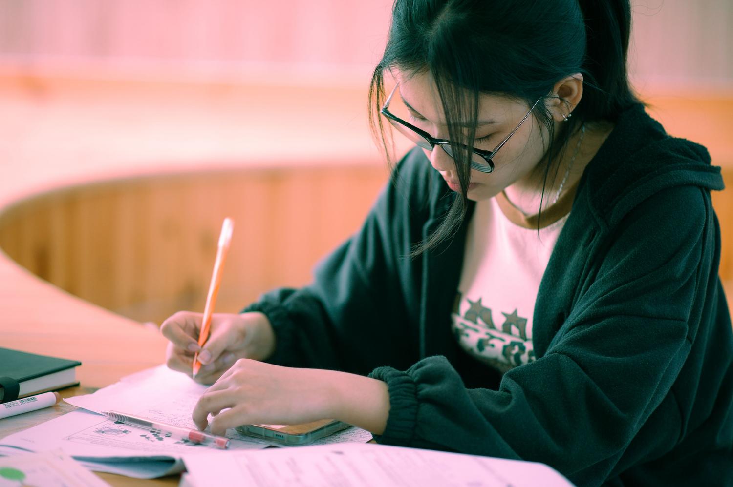 Mujer joven con gafas y sudadera totalmente inmersa en la escritura manual sobre papel, reflejando la conexión cognitiva del estudio analógico. Mujer joven con gafas y sudadera totalmente inmersa en la escritura manual sobre papel, reflejando la conexión cognitiva del estudio analógico.