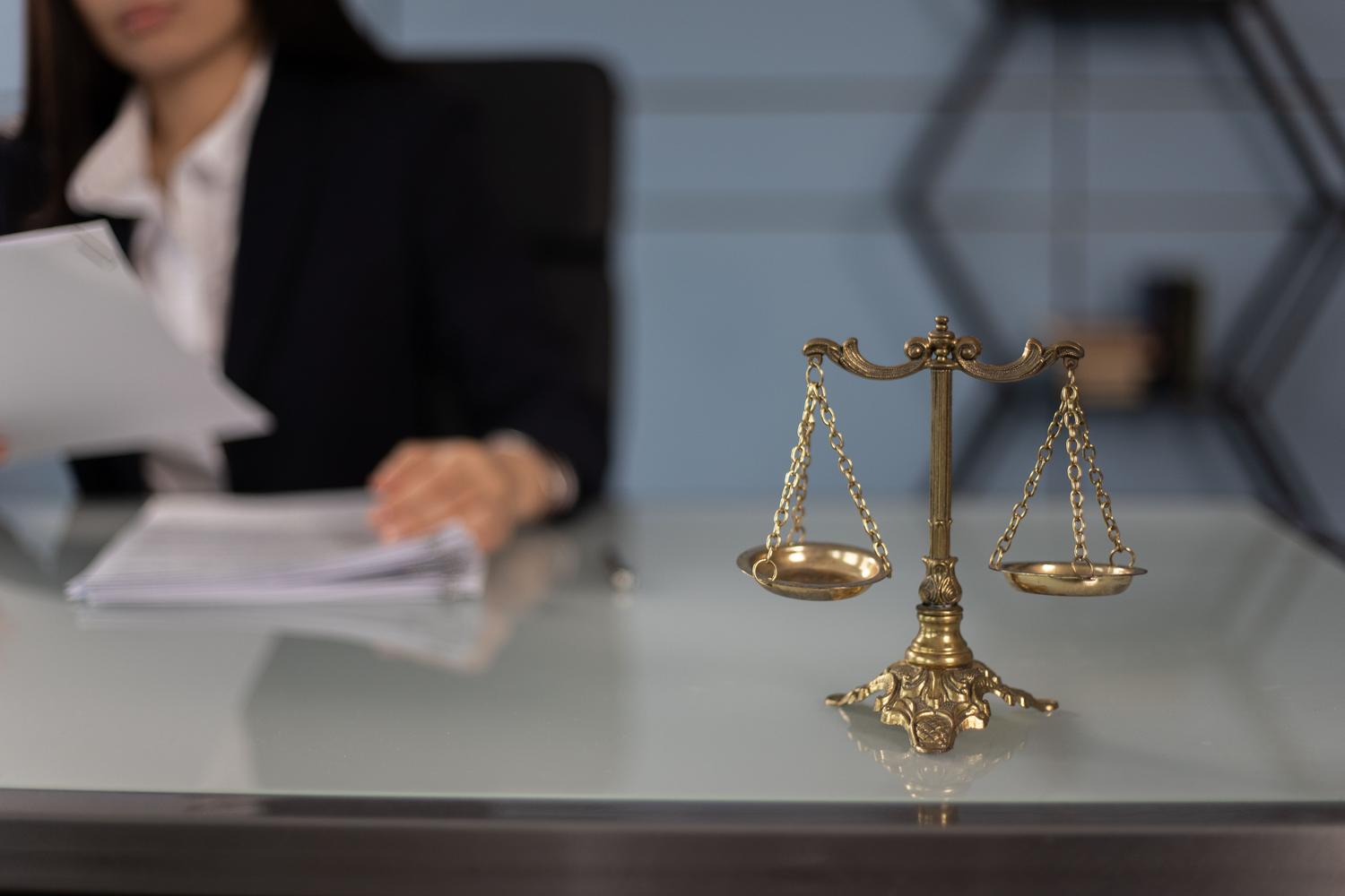 Balanza de la justicia en un escritorio profesional con un abogado desenfocado al fondo, aportando un contexto humano a la responsabilidad legal. Balanza de la justicia en un escritorio profesional con un abogado desenfocado al fondo, aportando un contexto humano a la responsabilidad legal.